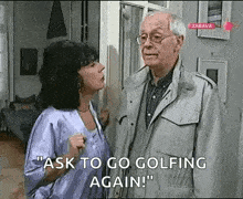 a man and a woman are standing next to each other in a living room . the woman is asking the man to go golfing again