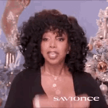 a woman with curly hair is standing in front of a christmas tree wearing a black top and necklace .