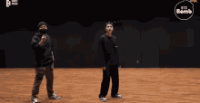 two men are standing next to each other on a wooden floor with a bts bomb logo in the background