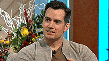 a man is sitting in front of a bunch of flowers and talking on a television show .