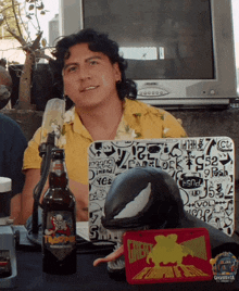 a man in a yellow shirt sits in front of a microphone with a sticker on his laptop that says carlock