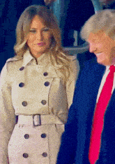 a man in a blue suit and red tie stands next to a woman in a trench coat