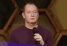 a man in a purple shirt is holding a microphone and making a gesture .