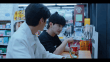 two young men are sitting at a table in a convenience store talking to each other