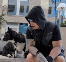 a man in a black hoodie sits next to a black and white dog in front of a building that says mentech