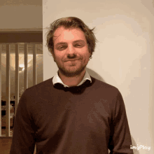 a man with a beard wearing a brown sweater and a white shirt is smiling in front of a wall