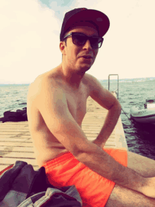 a shirtless man wearing sunglasses and a hat is sitting on a dock near the water