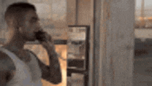 a man in a white tank top is talking on a cell phone in a room .