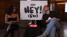 a man and woman sit in front of a sign that says hey
