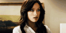 a woman with long dark hair is wearing a white shirt and looking at the camera with a surprised look on her face .