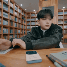 a young man sits at a table in front of a bookshelf with a cell phone on it