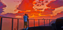 a couple standing on a balcony looking at a sunset with a car in the background