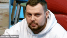 a man with a beard is wearing a white hoodie and a red chair