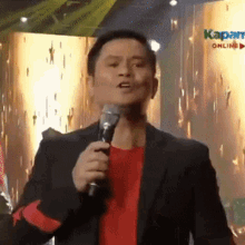 a man in a suit and red shirt is holding a microphone and speaking into it .