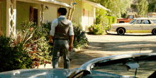 a man in a vest stands in front of a house with a yellow car parked in front of it