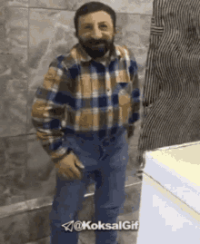 a man with a beard wearing a plaid shirt and jeans is standing in front of a refrigerator .