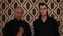 two men standing in front of a patterned wallpaper