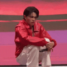 a man in a red jacket and white pants is sitting on a stage with his legs crossed and holding a microphone .