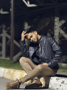 a man wearing a leather jacket sits on a curb with his hand on his head