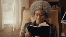 an elderly woman with glasses is sitting in a chair reading a book .