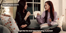 two women are sitting on a couch talking to each other and one of them is holding a purse