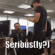 a man standing next to a man sitting at a desk with the words serious ( ly ) written on it