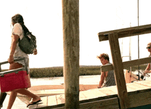 a woman is carrying a cooler on a dock next to a man sitting on a boat .