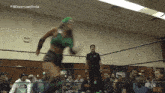 a woman in a green top and black shorts is jumping in a wrestling ring while a referee watches