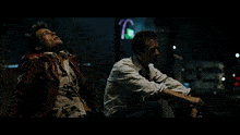 a man in a red jacket sits next to another man in a white shirt in front of a neon sign that says tc