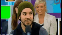 a man wearing a beanie is talking into a microphone while a woman watches .