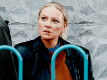 a woman wearing a brown sweater and a black jacket is standing in front of a blue fence