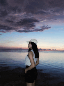 a woman wearing a white hat stands in front of the ocean at sunset