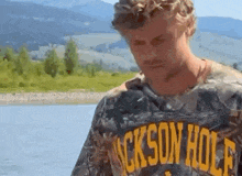 a man wearing a jackson hole t-shirt is standing in front of a lake .