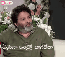 a man with a beard and a green shirt is sitting on a couch in front of a bunch of flowers .