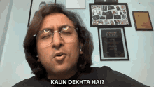 a man wearing glasses says " kaun dekhta hai " in front of a wall of framed pictures