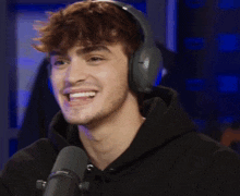 a young man wearing headphones and a black hoodie smiles in front of a microphone
