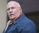 a bald man with glasses and a mustache wears a blue jacket