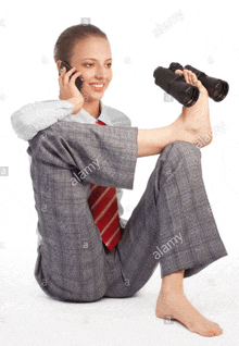 a woman in a suit and tie is sitting on the floor talking on a cell phone and holding binoculars