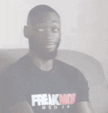 a man wearing a black t-shirt that says freakmob media is sitting on a couch .