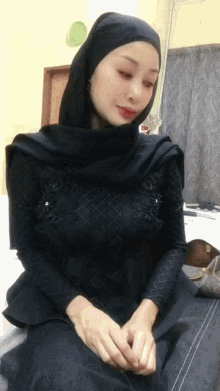 a woman wearing a black hijab sits on a bed with her hands folded