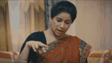 a woman in a red saree is sitting on a couch and looking at something .