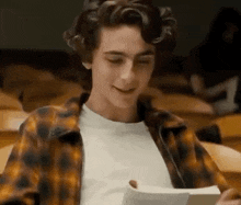 a young man in a plaid shirt is sitting in a lecture hall reading a book .