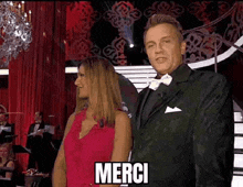 a man in a suit and bow tie is standing next to a woman in a red dress who is saying merci