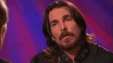 a man with long hair and a beard is talking to another man while wearing a leather jacket .