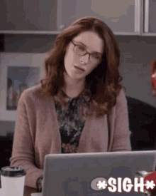 a woman wearing glasses sits at a desk with a laptop and a sign that says sigh