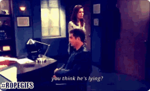 a man sitting at a desk with a woman standing behind him asking if he 's lying