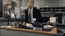 a man in a suit and tie is standing in front of a desk with a computer and a sign that says alice me .
