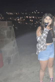 a girl in shorts and a plaid shirt holds a cell phone