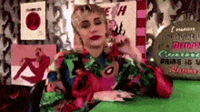 a woman in a colorful shirt is sitting at a table with a sign that says i mean i 'm over it