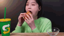 a woman in a green sweater is eating a sandwich next to a subway cup